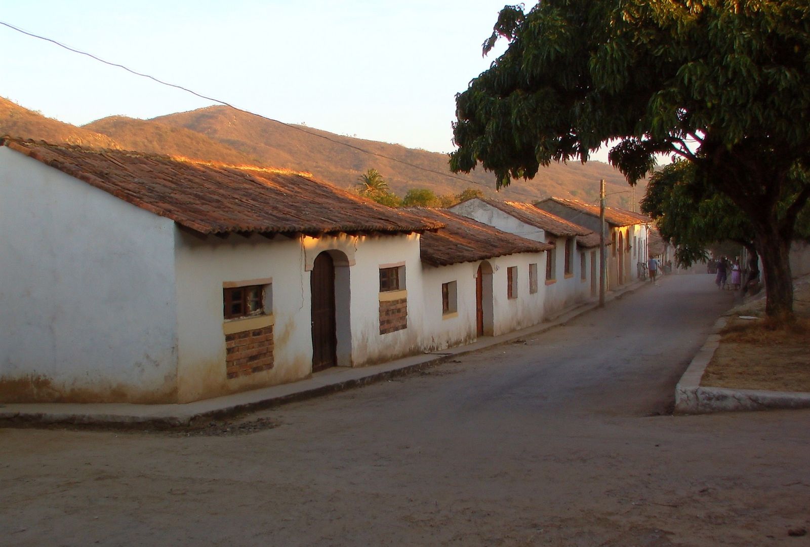 Calle ancha y polvorienta de un pueblo de calor seco, con casas bajas de adobe blanco y techo de teja y cerros pelados al fondo