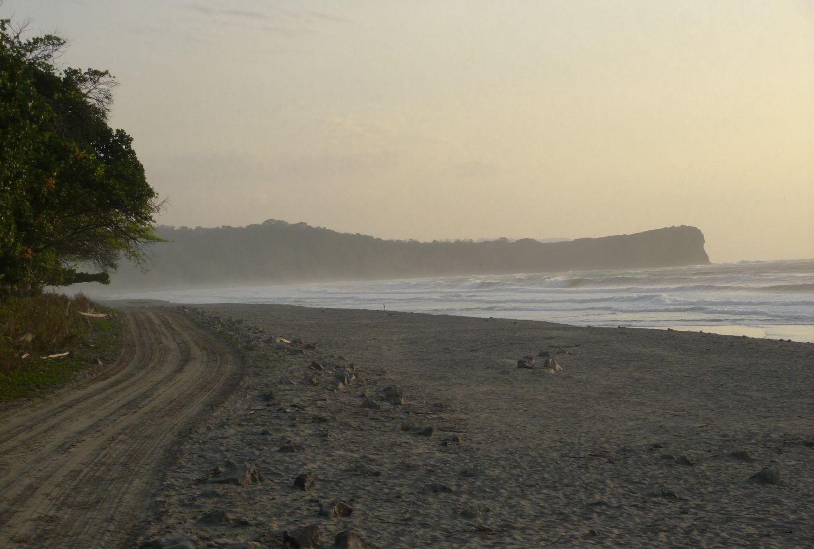Camino de lastre paralelo a una playa larga de oleaje constante, con el monte seco de un lado y bruma marina al atardecer