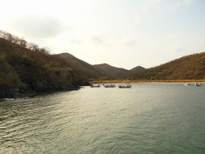 Bahía de arena gris con lanchas de pesca fondeadas cerca de la orilla y cerros secos cerrando los dos extremos