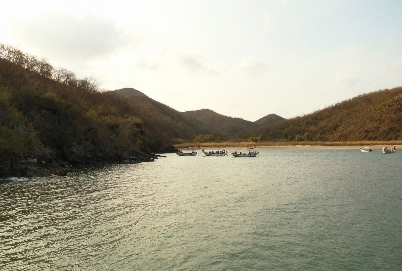 Bahía de arena gris con lanchas de pesca fondeadas cerca de la orilla y cerros secos cerrando los dos extremos