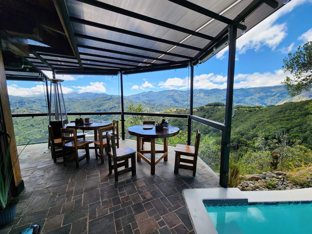 Mesas redondas de madera en la terraza techada, junto al borde de la piscina y con vista a las montañas