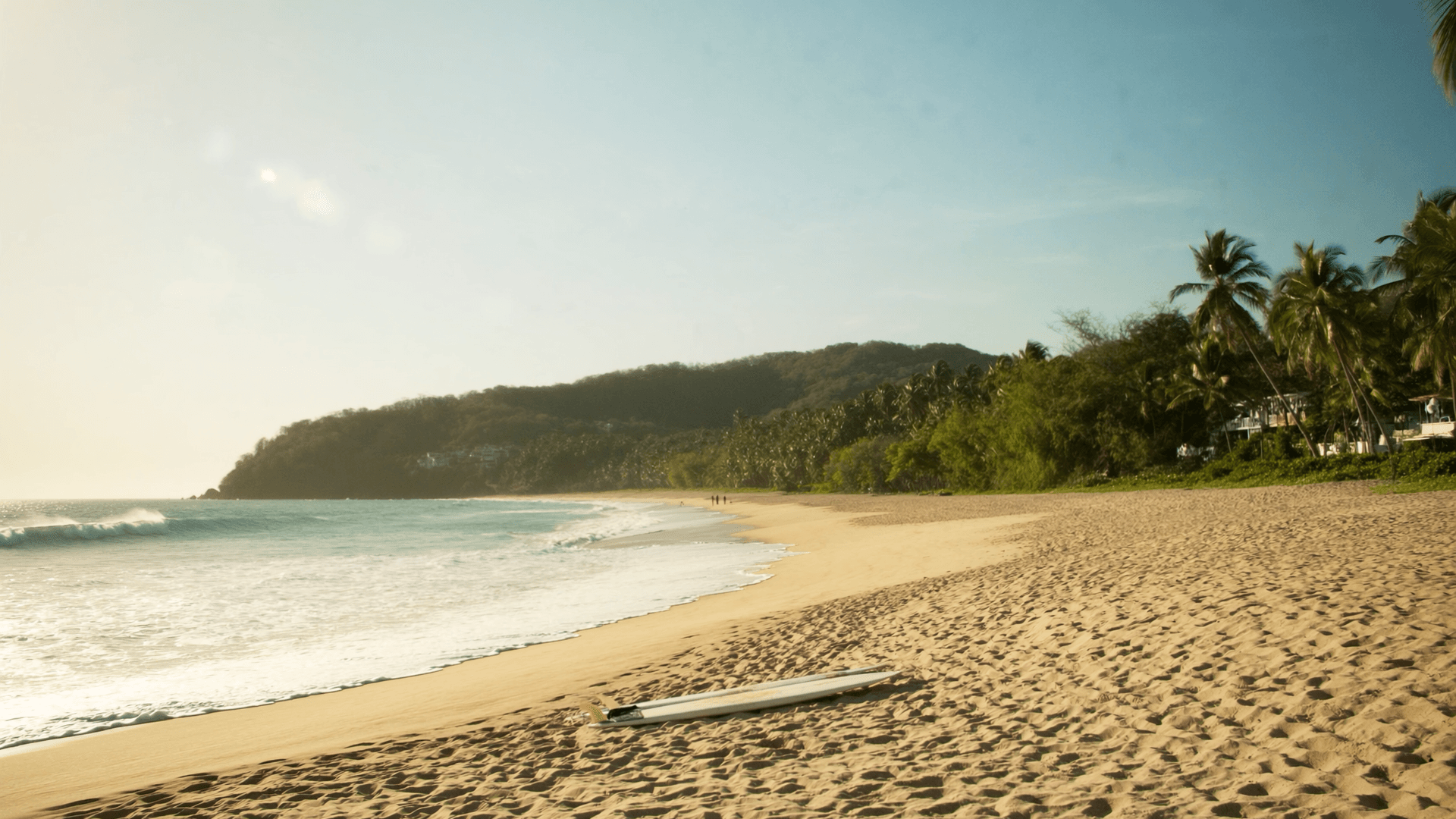 Tabla de surf sobre la arena de una playa de Tamarindo al atardecer, con palmeras y un cerro boscoso al fondo